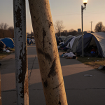 Worn baseball bat leaning against metal fence at dusk with golden light and flickering streetlight over a homeless encampment