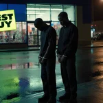 Suspects stand on wet sidewalk with handcuffs in front of neon‑lit Best Buy storefront and security guard behind them.