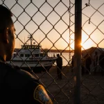 Border patrol agent standing behind fence watching patrol boat dock with sunset glow on river and migrants in distance.