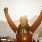 Camille Rast raising her arms on podium with hopeful glow blurred mountain silhouette in background