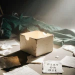 A small glowing box sits on the floor with crumpled papers and broken game rule cards scattered around and a dark track suit