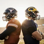 Two college football quarterbacks stand back-to-back on a sunlit gridiron with helmets and crowd goalposts blurred behind the