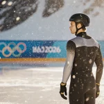 Ilia Malinin stands on a frozen pond gazing at the Alps while a 2026 Olympics logo floats on the ice warm glow highlights his
