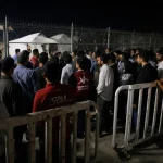 Aid workers stand behind barricades while Gaza clinic and waiting people appear through fence at border crossing.