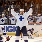 Aron Kiviharju hoists the victory puck with Finnish teammates celebrating while the defeated American team slumps in warm lig