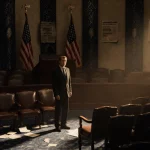 Lone lawmaker standing beside empty seat with dim House of Representatives chamber and golden light and faded flag