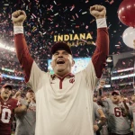 Coach Curt Cignetti triumphantly raising his arms with cheering Indiana fans in red and cream and a confetti-filled stadium