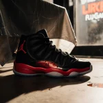 Jordan 12 sneakers resting on polished concrete floor with cracked vinyl tablecloth and blurred restaurant sign reflection.