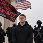 Judge standing outside courthouse with officers in tactical gear and a flag and protest banner with red letters