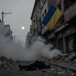 Person lies on the sidewalk with smoke and rubble scattered around and a Ukrainian flag waving above a shattered storefront