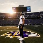 Lamar Jackson stands alone at midfield looking up at sunset sky with Steelers logo behind him and fading Ravens colors seats.