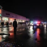 Lone figure standing near the entrance of Suite B148 with neon strip mall and police cars reflecting off wet pavement