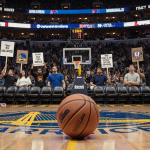 Fans holding protest signs with an abandoned basketball on the court and the Warriors logo on the arena wall.