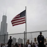 American flag rises with chain-link fence and police guard near Chicago skyline with protesters chanting.