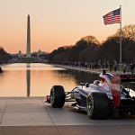 Sleek Formula 1 car parked with sunset orange sky and spectators looking at phones.