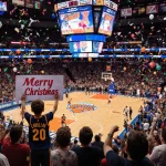 Boy in Cavaliers jersey holding Merry Christmas sign with confetti and balloons amid packed Knicks arena