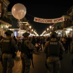 National Guard troops stand along Bourbon Street with streetlights illuminating uniforms and a faint memorial in background.