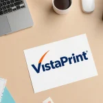 Desk organized with VistaPrint logo and laptop and printer and a postcard and promo code folder on warm beige background