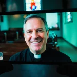 Smartphone screen showing Pastor Mike Schmitz speaking to congregation with distorted blue‑green glow and church background.