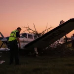Police officer examining plane wreck with wing tangled in branches and passengers escorted by staff under orange-pink dusk sk