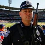 Police officer standing at attention with rifle near Sydney Cricket Ground while fans hold Australia vs England banners