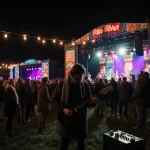 Musician plays darkwave guitar on colorful festival stage with string lights and crowd bundled in coats watching