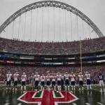 Alabama players standing and gazing up with a rain‑soaked Rose Bowl archway shadowing the field