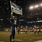 Seahawks player holding championship flag aloft with golden stadium lights and scattered confetti on empty Lumen Field.