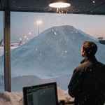 Air traffic controller standing under flickering light with massive snowdrift engulfing terminal entrance at dusk
