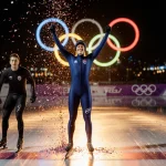 Casey Dawson triumphantly raising arms at speed skating finish line with confetti and golden light and Olympic rings behind