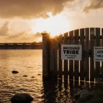 Wooden gate with Miccosukee Tribe and Rep. Lauren Boebert signs stands in sunset water with faint water treatment plant.