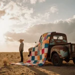 Lone figure standing under a patchwork quilt over a rusty truck with warm sunlit dry Texas landscape and storm clouds.