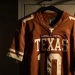 Worn Texas Longhorns jersey hangs on locker with warm golden light and subtle seat grid backdrop.