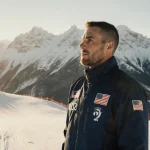 Tim Burke a biathlete standing at the edge of a ski slope with mountains and a golden glow and an American flag pin