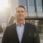 Urban Meyer standing confidently with modern campus building and protest sign in background light casting warm glow