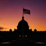 Venezuelan Presidential Palace rises under a sunset sky with an American flag on dais and barbed wire surrounding the buildin