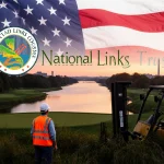 Maintenance worker in orange vest standing with forklift beside golf course at sunset over Potomac River and flag.