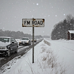 Cars moving along a rural road with frosty wipers and snow‑covered tires under a gray winter sky and a weathered FM Road sign